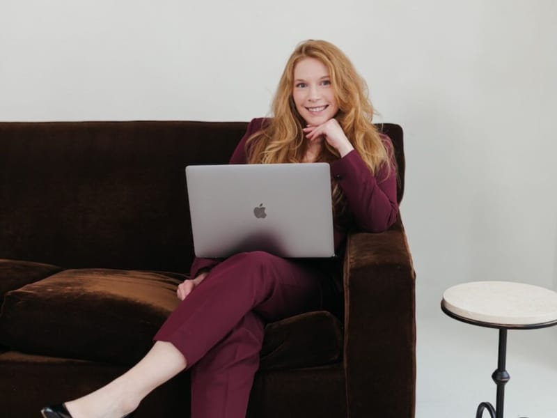 Cheryl Ann seated on a dark brown velvet sofa with a MacBook laptop, smiling at camera, wearing a burgundy suit